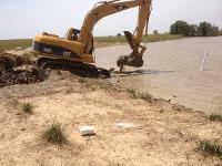 The excavator is at the edge of the pond, digging the trench into the pond itself.