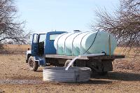 This photo shows a view of a water tank truck pumping water into a metal livestock watering tank.