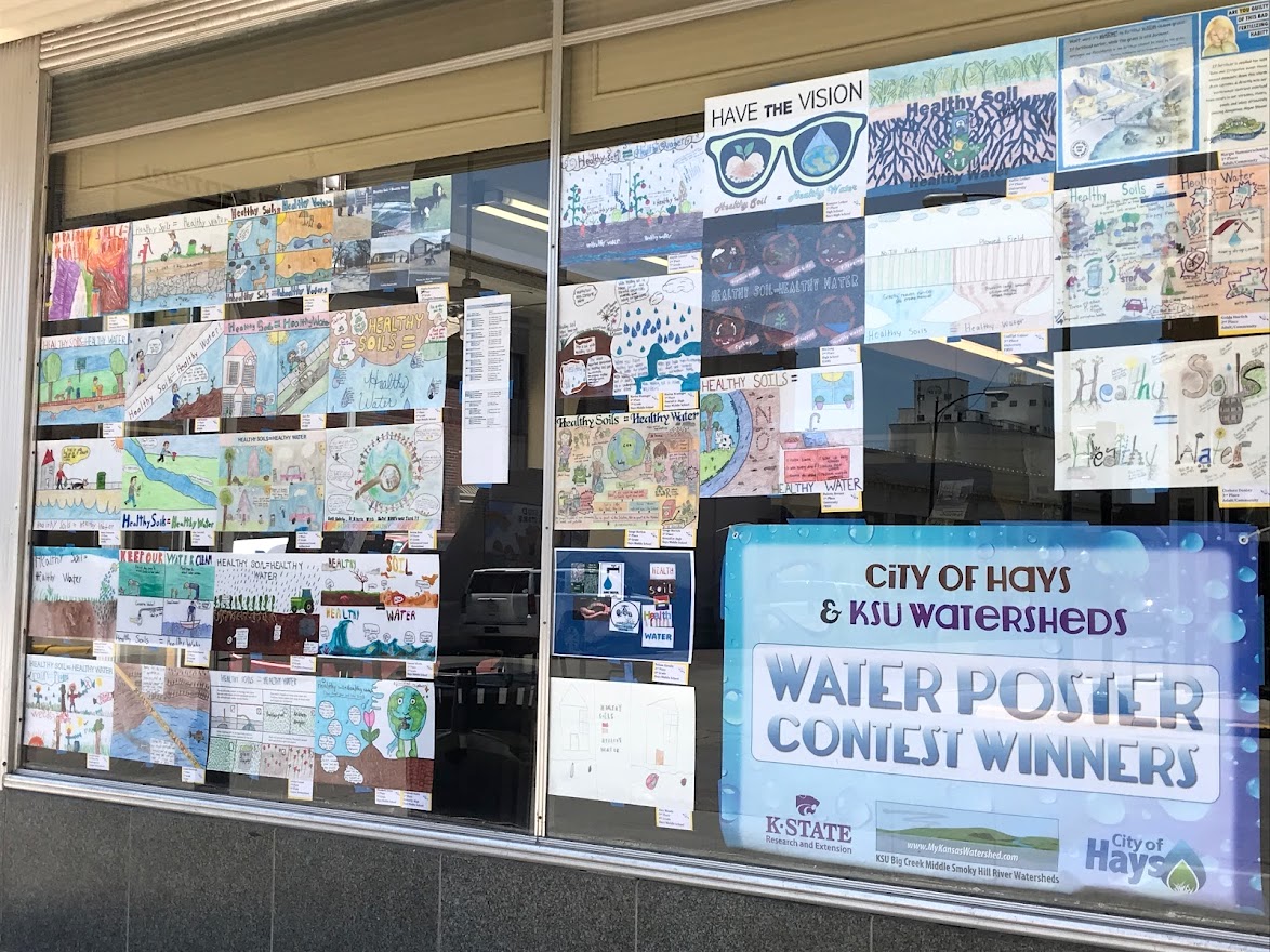 Rows of hand-drawn posters with the theme Healthy Soils, Healthy Water are displayed in the window of a local business in Hays, Kansas