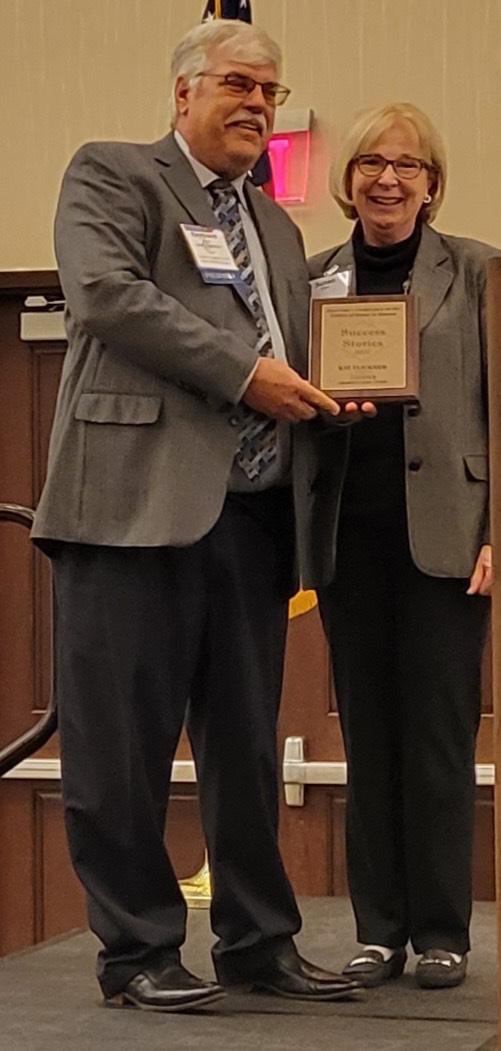 Ray Flickner, left, wears a grey suitjacket and holds a small plaque in front of Susan Flickner, right, who smiles at the camera.