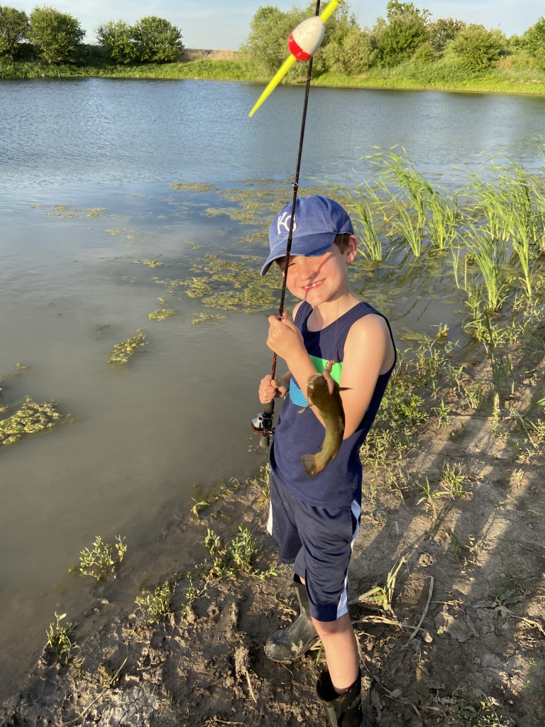 A boy, wearing a hat and holding a fishing rod, shows the fish he caught with a muddy pond in the background.