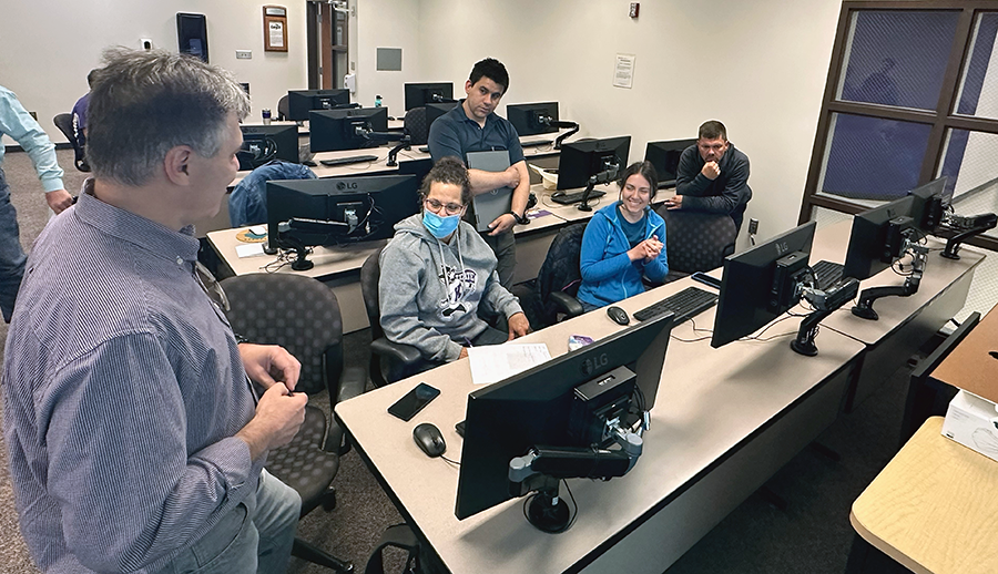 A man in a striped shirt stands in the foreground, speaking with a group of adult students sitting at a table with black-colored computer monitors.