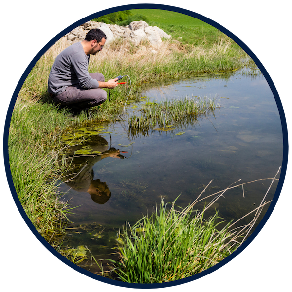 A man kneels by the side of a still pond and looks at a measuring instrument that he has placed in the water