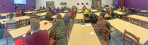 People sit at tables in a large room, listening to a presentation