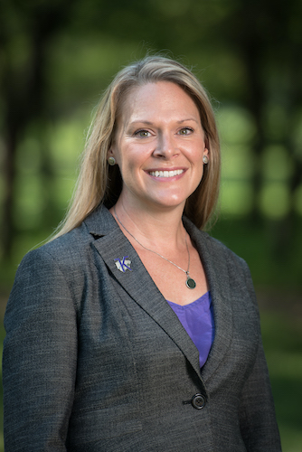 Susan Metzger, with long blonde hair, a grey suit jacket and purple shirt, smiles at the camera