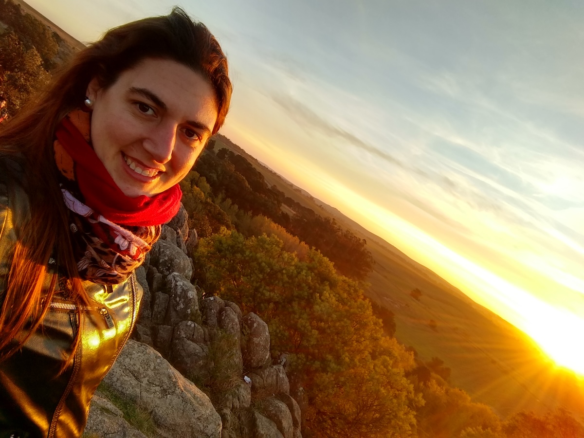 A young woman with dark hair, and wearing a jacket and a bright red scarf, smiles into the camera in front of the setting sun