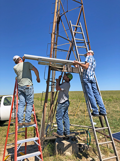 Three men in blue jeans, two standing on ladders and the third balanced on a cross beam, hold a large metal solar panel parallel to the ground.